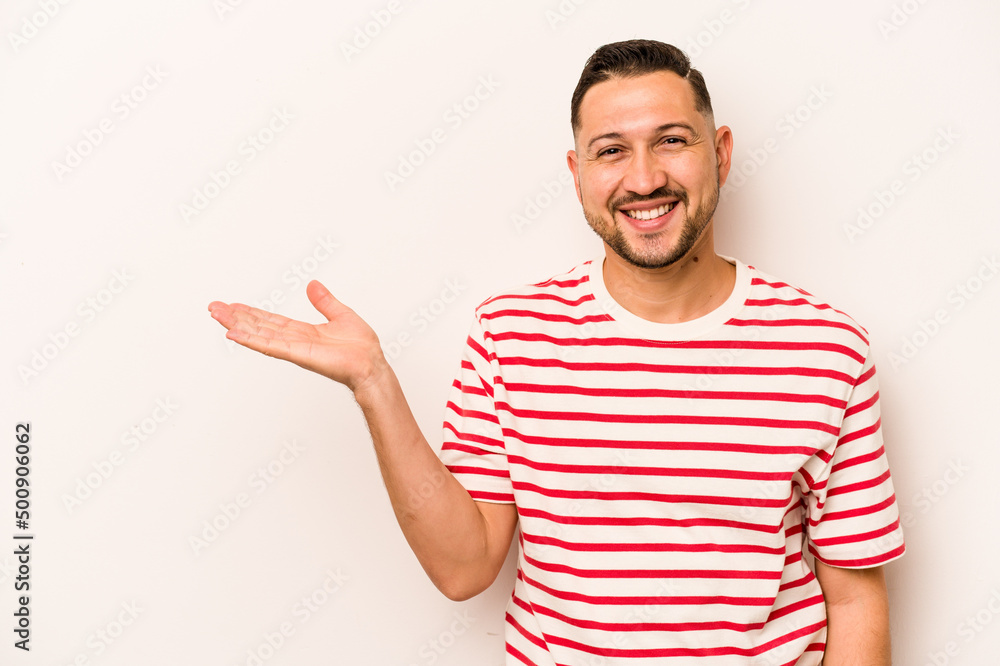 Young hispanic man isolated on white background showing a copy space on a palm and holding another hand on waist. Young hispanic man isolated on white background showing a copy space on a palm and holding another hand on waist.