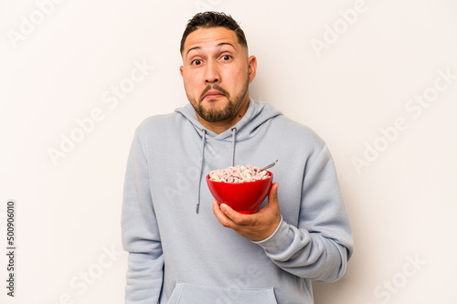 Hispanic man eating cereals isolated on white background shrugs shoulders and open eyes confused.