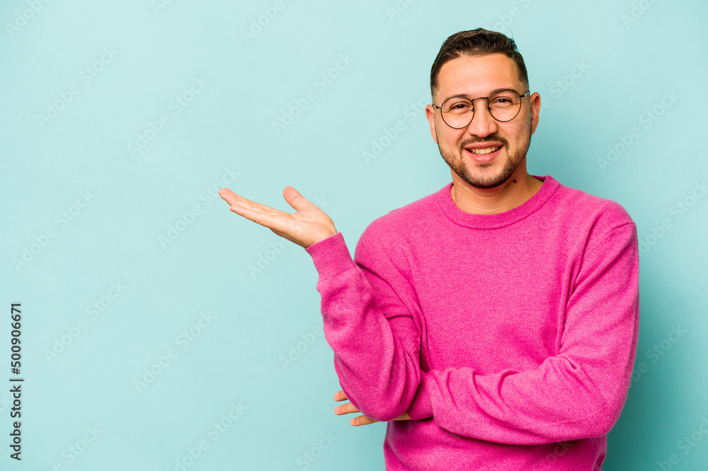 Fototapeta premium Young hispanic man isolated on blue background showing a copy space on a palm and holding another hand on waist.