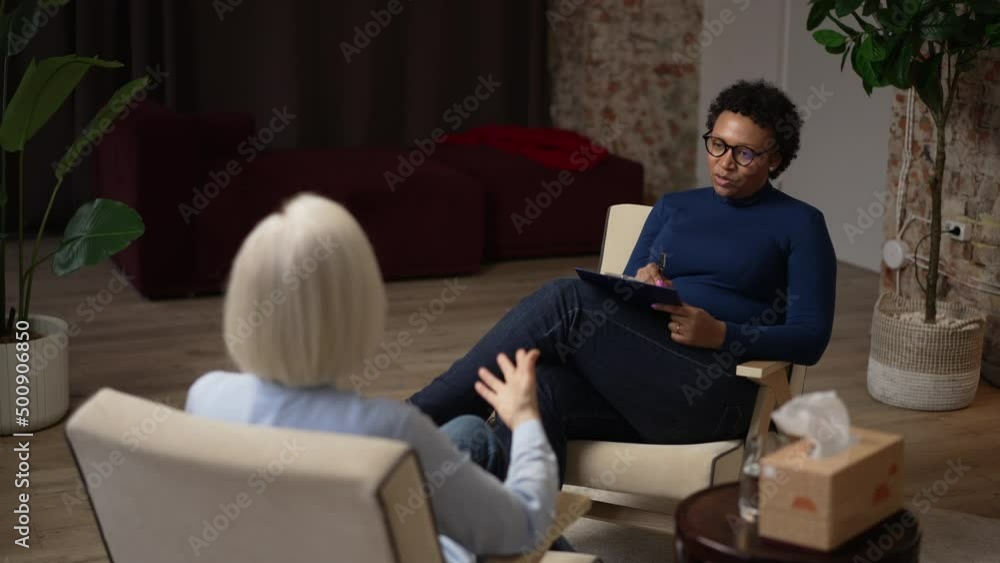 Experienced psychoanalyst is consulting female patient and sitting in chair in modern office spbas. 4k Young African American woman talks to mature person and looks with smile, writes on clipboard and Experienced psychoanalyst is consulting female patient and sitting in chair in modern office spbas. 4k Young African American woman talks to mature person and looks with smile, writes on clipboard and