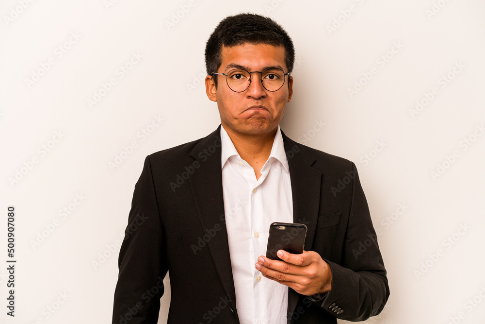 Young business hispanic man holding mobile phone isolated on white background shrugs shoulders and open eyes confused. Young business hispanic man holding mobile phone isolated on white background shrugs shoulders and open eyes confused.