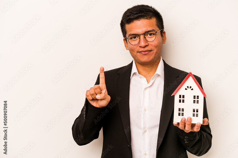 Young business hispanic man holding a little house isolated on white background showing number one with finger. Young business hispanic man holding a little house isolated on white background showing number one with finger.