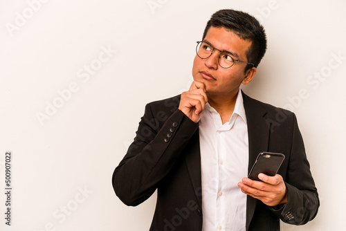 Fotografie Young business hispanic man holding mobile phone isolated on white background looking sideways with doubtful and skeptical expression