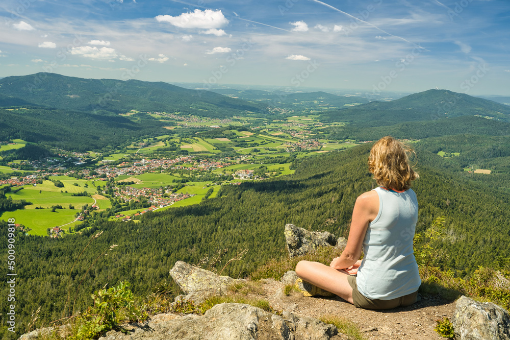 A woman sitting on mount Osser, overlooking the landscape of Lamer Winkel with the small town Lam in the Bavarian Forest