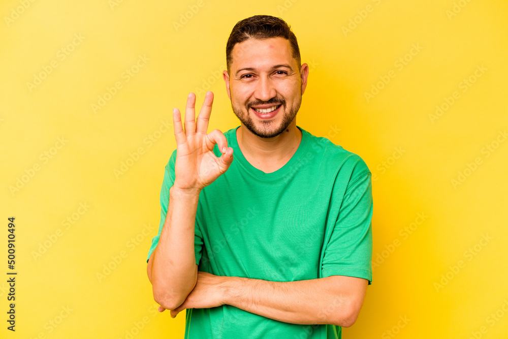 Young hispanic man isolated on yellow background winks an eye and holds an okay gesture with hand. Young hispanic man isolated on yellow background winks an eye and holds an okay gesture with hand.
