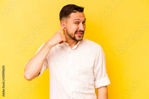 Young hispanic man isolated on yellow background covering ears with fingers, stressed and desperate by a loudly ambient. © Asier