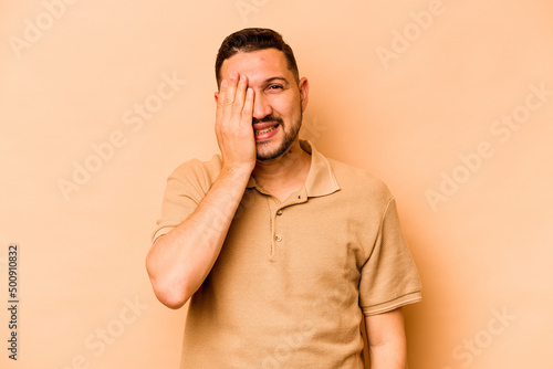 Young hispanic man isolated on beige background having fun covering half of face with palm.