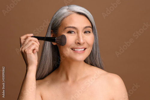 Lovely middle-aged Asian woman with long gray hair holding blush brush near face standing isolated on brown, korean female applying makeup, using powder for her skin type