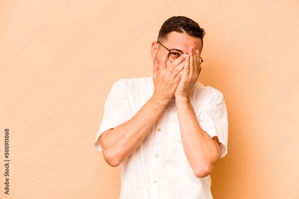 Young hispanic man isolated on beige background blink at the camera through fingers, embarrassed covering face. Young hispanic man isolated on beige background blink at the camera through fingers, embarrassed covering face.