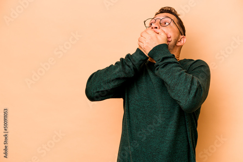 Young hispanic man isolated on beige background thoughtful looking to a copy space covering mouth with hand.