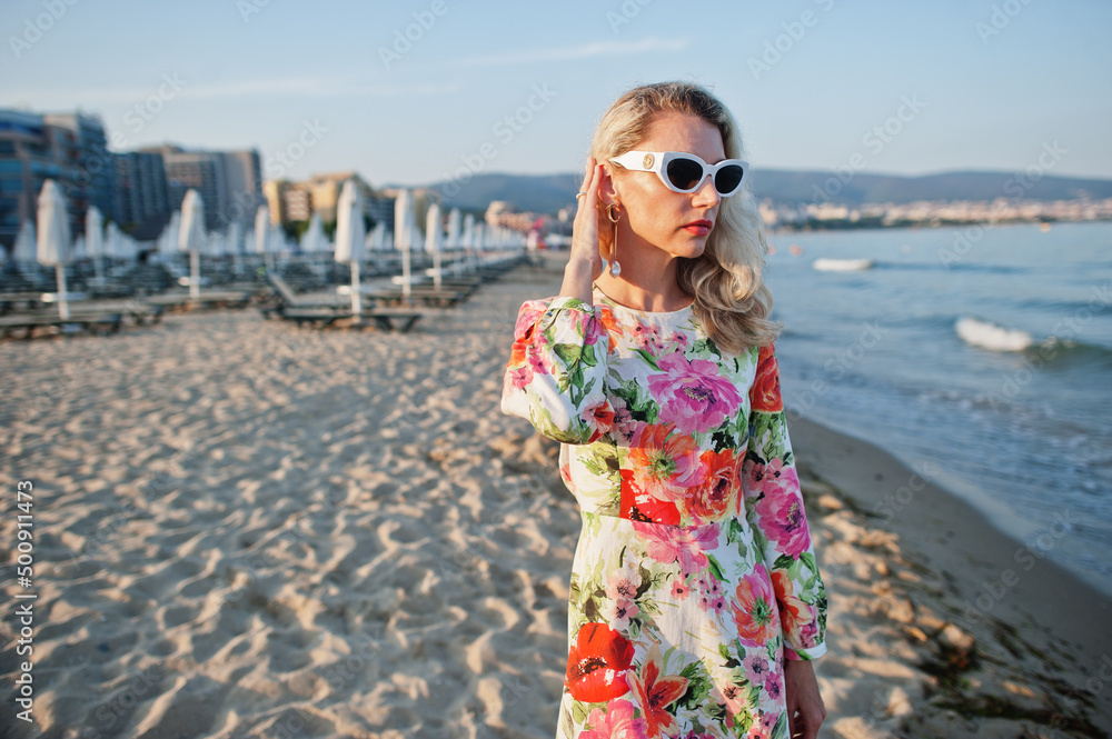 Blonde woman wearing dress and sunglasses standing on sand beach at sea shore enjoying view of sunset.