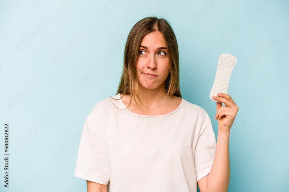 Young caucasian woman holding sanitary napkin isolated on blue background confused, feels doubtful and unsure. Young caucasian woman holding sanitary napkin isolated on blue background confused, feels doubtful and unsure.