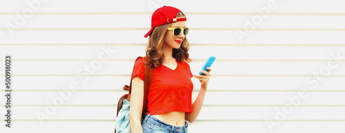 Portrait of modern young woman with smartphone wearing red baseball cap, backpack on white background, blank copy space for advertising text © rohappy