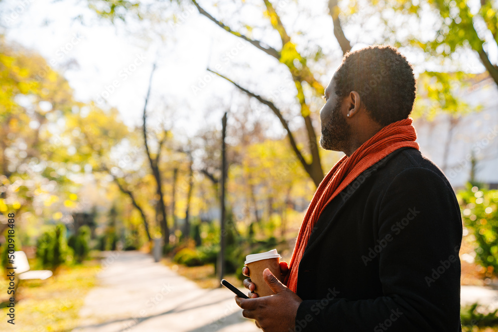 Black man using cellphone and drinking coffee while strolling in park Black man using cellphone and drinking coffee while strolling in park