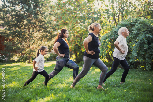Four females of different generations of family doing physical exercises for stretching legs together on backyard full of green grass and trees. Spending time together. 