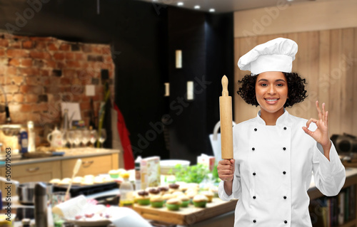 cooking, culinary and people concept - happy smiling female chef in toque or baker with rolling pin over restaurant kitchen background