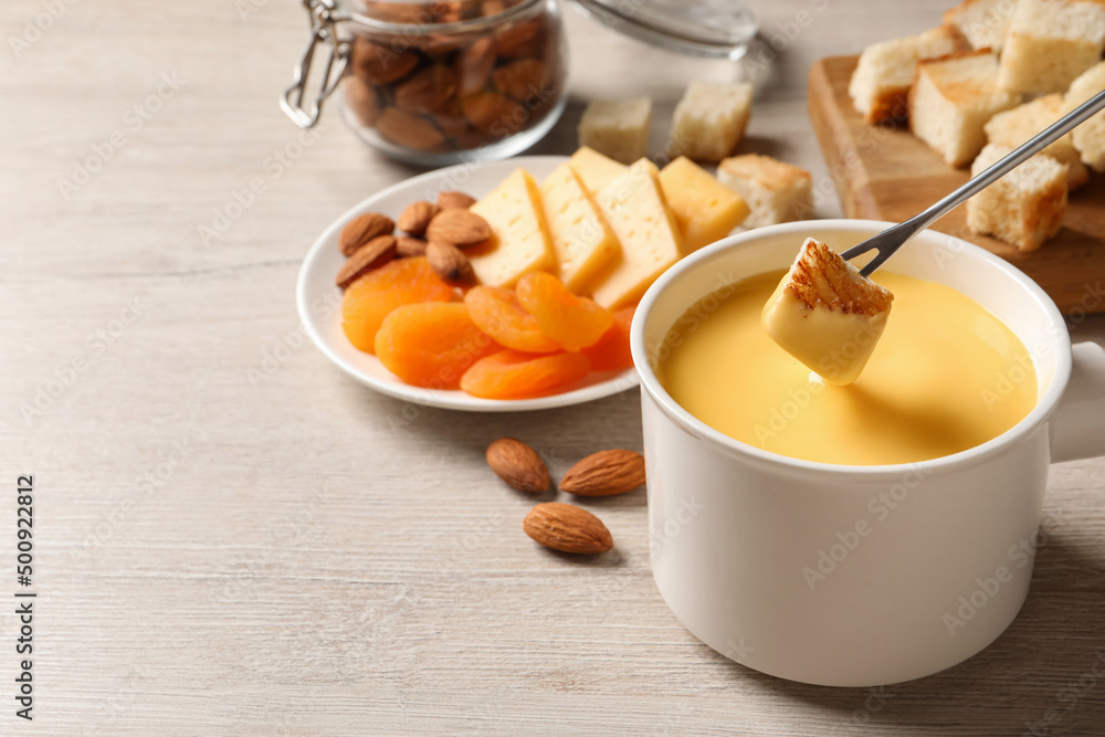Dipping piece of bread into tasty cheese fondue on light wooden table, space for text Dipping piece of bread into tasty cheese fondue on light wooden table, space for text