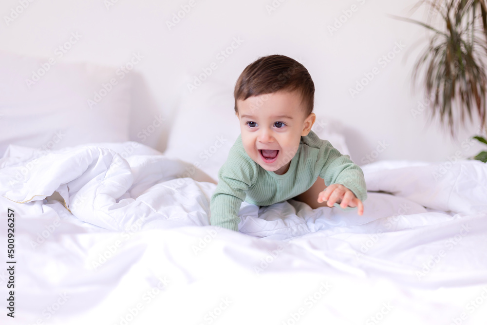 cheerful baby in a green cotton bodysuit is lying on his stomach on a white bed cheerful baby in a green cotton bodysuit is lying on his stomach on a white bed