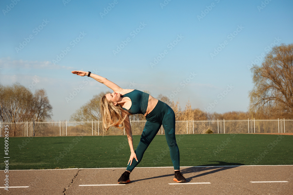 Athletic woman is stretching her arm and side outdoors at the stadium.