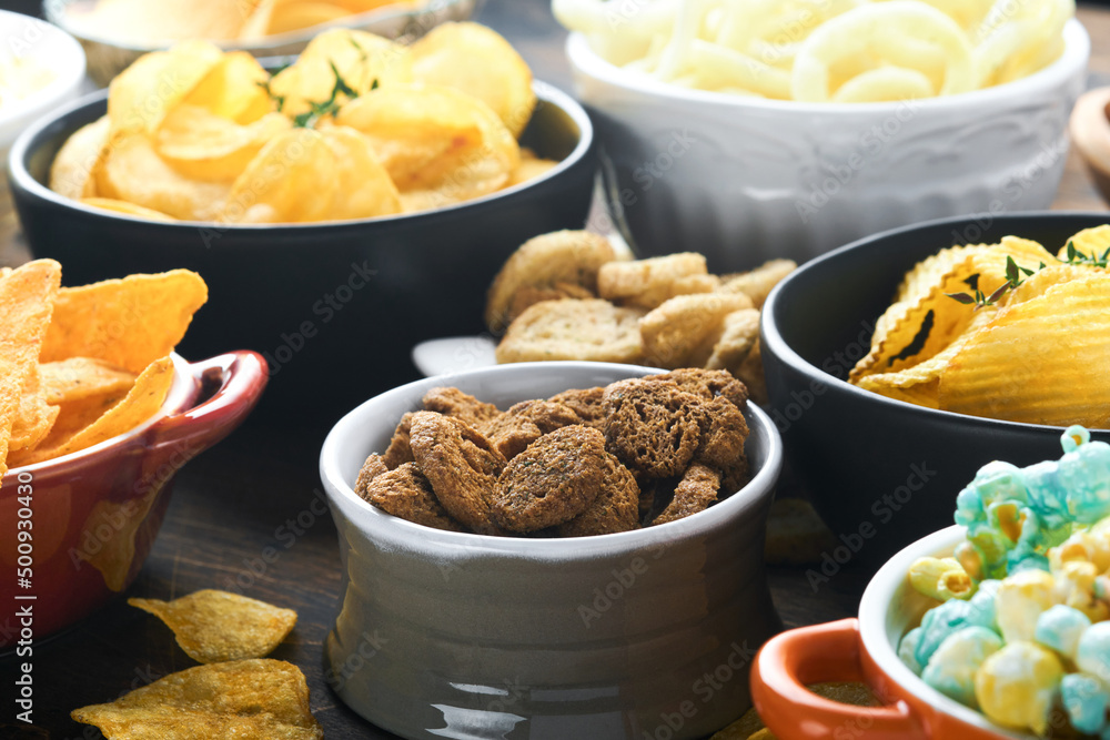 Unhealthy food or snacks. All classic potato snacks with peanuts, popcorn and onion rings and salted pretzels in bowl plates on old wooden background. Unhealthy food for figure, heart, skin, teeth. Unhealthy food or snacks. All classic potato snacks with peanuts, popcorn and onion rings and salted pretzels in bowl plates on old wooden background. Unhealthy food for figure, heart, skin, teeth.