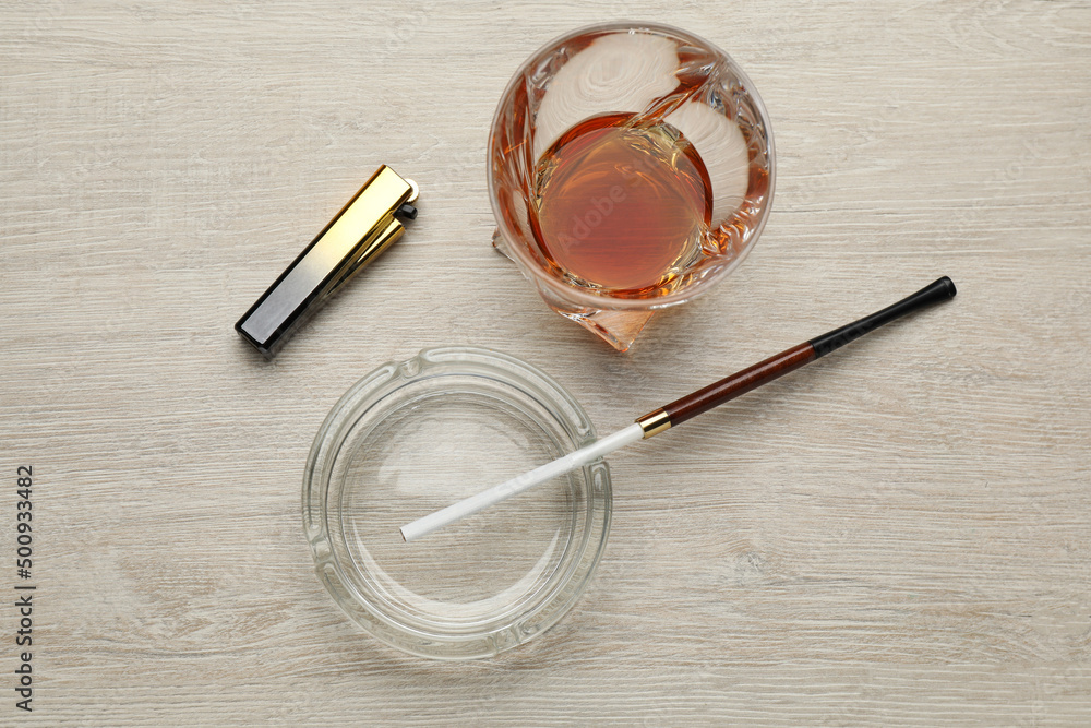 Glass ashtray with long cigarettes holder, lighter and alcohol drink on wooden table, flat lay Glass ashtray with long cigarettes holder, lighter and alcohol drink on wooden table, flat lay