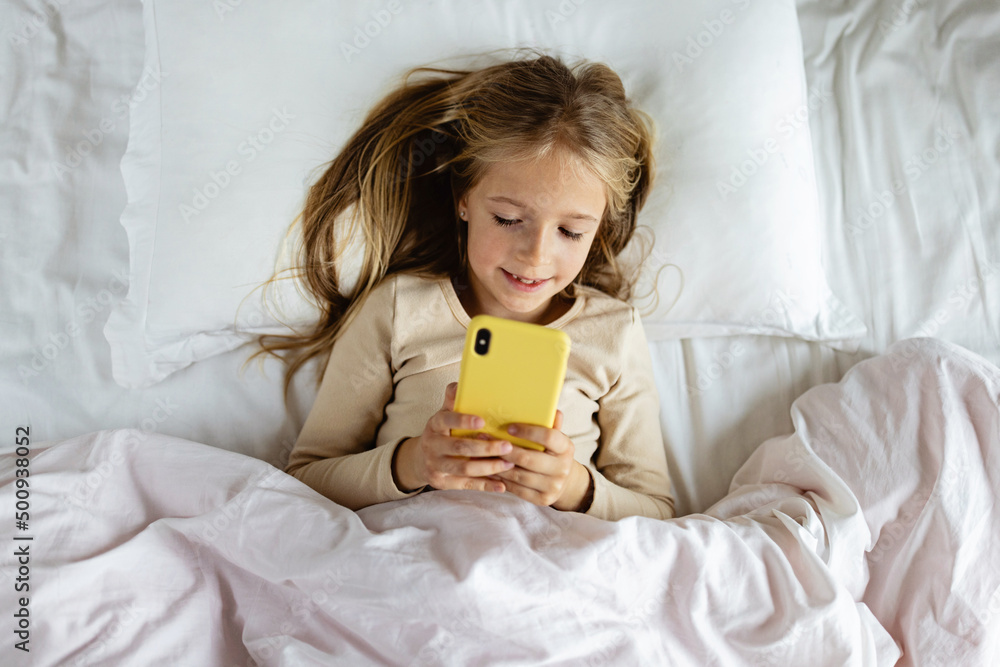 Cute little caucasian girl with blonde hair in pajamas at home during coronavirus pandemic quarantine and using mobile phone. Stay at home during covid-19 lockdown concept.
