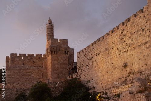 Twilight view of the landmark, stone Tower of David minaret as seen through branches and leaves of trees along the golden sunset walls of the Old City of Jerusalem.