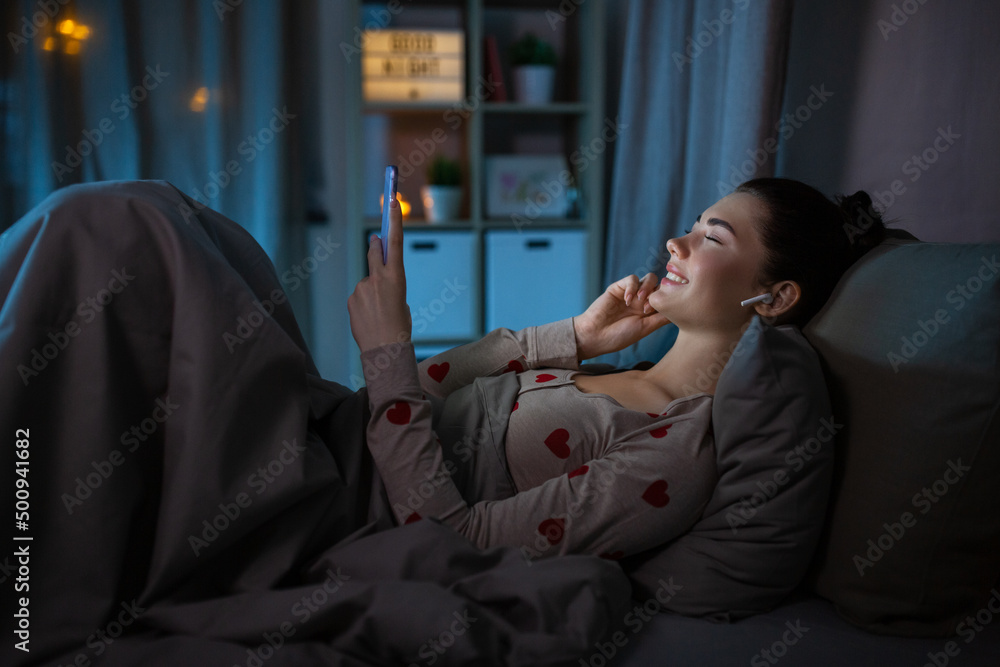 technology, bedtime and people concept - happy teenage girl with smartphone and earphones enjoying music lying in bed at home at night