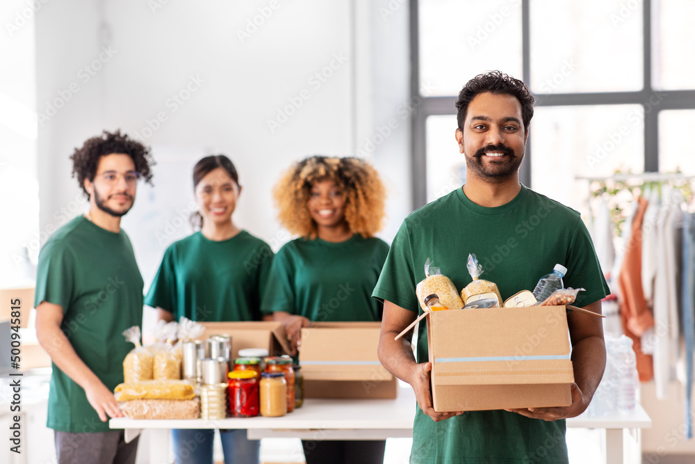 charity, donation and volunteering concept - happy smiling male volunteer with food in box and international group of people at distribution or refugee assistance center charity, donation and volunteering concept - happy smiling male volunteer with food in box and international group of people at distribution or refugee assistance center