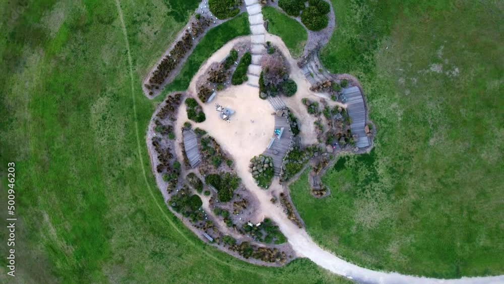 Drone looking down with a slow spin. A green field with a path passing through the shot with a sandy round middle with shrubs in a nice pattern around it. Drone looking down with a slow spin. A green field with a path passing through the shot with a sandy round middle with shrubs in a nice pattern around it.