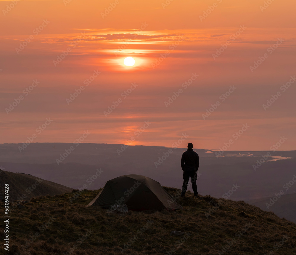 Silhouette of an adventurer and wild camping tent high in the mountains at sunset