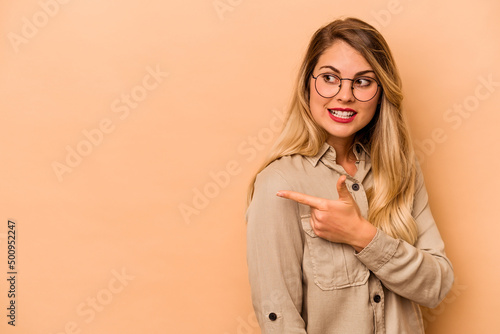 Young caucasian woman isolated on beige background points with thumb finger away, laughing and carefree.