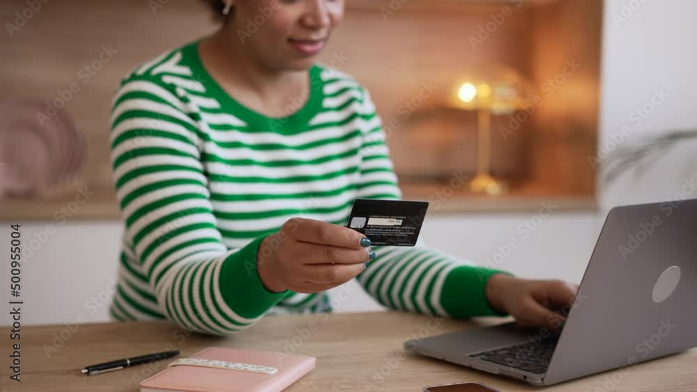 Young woman is holding card and doing shopping at table with laptop in home room spbd. 4k Close view of American African female looks with smile and types on computer, buys online with credit card in Young woman is holding card and doing shopping at table with laptop in home room spbd. 4k Close view of American African female looks with smile and types on computer, buys online with credit card in