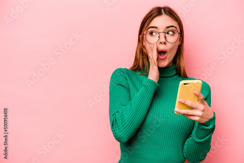 Young caucasian woman holding mobile phone isolated on pink background is saying a secret hot braking news and looking aside photo