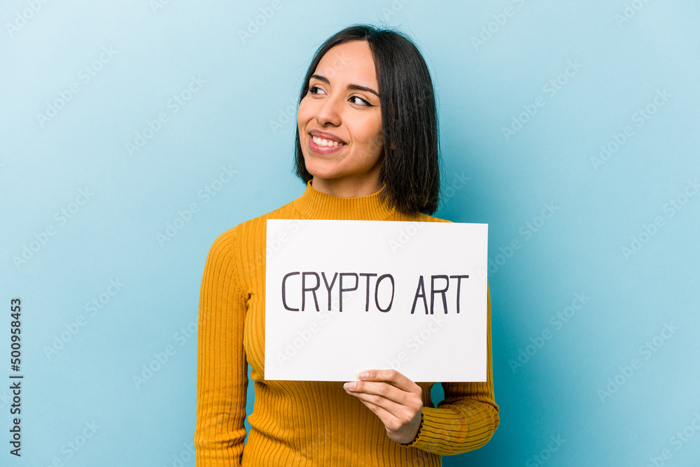 Young hispanic woman holding crypto art placard isolated on blue background looks aside smiling, cheerful and pleasant. Young hispanic woman holding crypto art placard isolated on blue background looks aside smiling, cheerful and pleasant.
