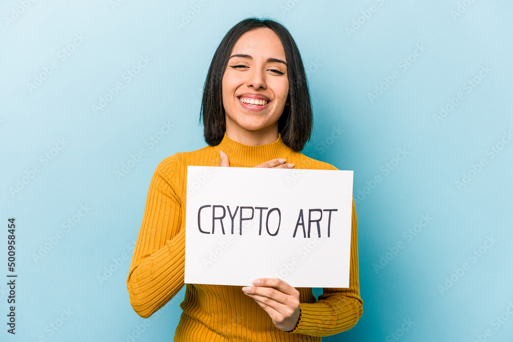 Young hispanic woman holding crypto art placard isolated on blue background laughs out loudly keeping hand on chest. Young hispanic woman holding crypto art placard isolated on blue background laughs out loudly keeping hand on chest.