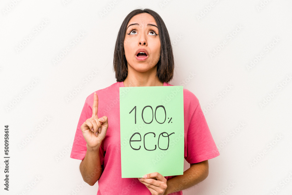 Young hispanic woman holding 100% eco placard isolated on white background pointing upside with opened mouth. Young hispanic woman holding 100% eco placard isolated on white background pointing upside with opened mouth.