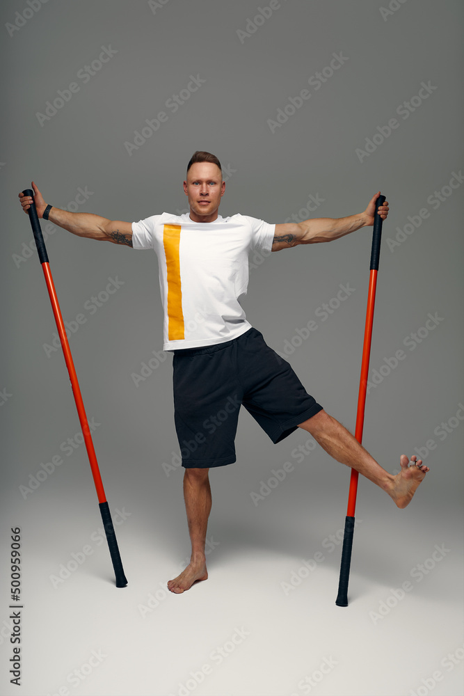 A fighter in white t-shirt and black shorts, stands in a fighting artificially pose and holds a fight stick on studio background.