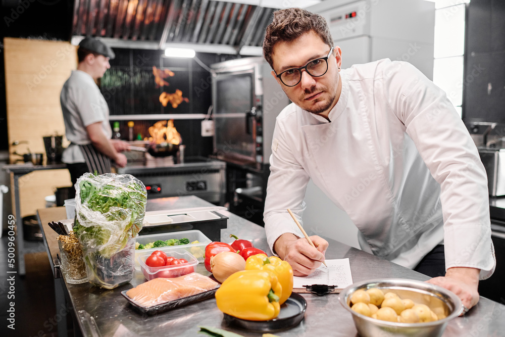 Portrait of young professional cook in uniform looking at camera while making list of food ingredients for dish in commercial kitchen