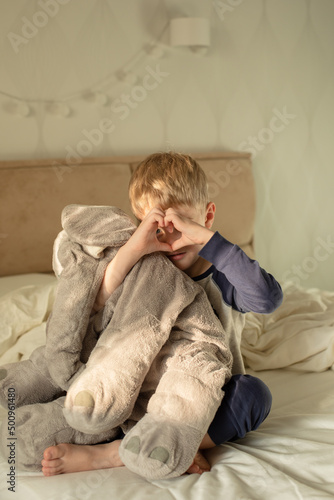 A little boy shows a heart with his hands sitting on the bed in the morning