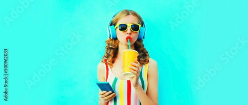 Summer colorful portrait of stylish happy young woman listening to music in headphones looking at smartphone and drinking fresh juice on blue background