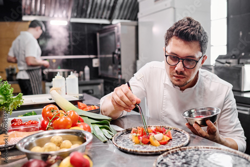Young chef in eyeglasses decorating dish with vegetables on plate at table in kitchen