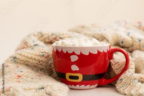 A christmas cup in the shape of a Santa Claus with cocoa and marshmallow stands next to a knitted sweater. Concept New Year, holidays, comfort