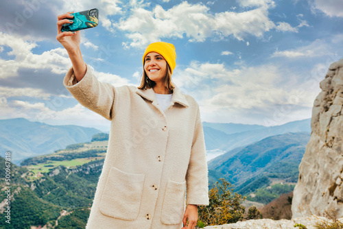 Portrait of young smiling stylish woman wears beige jacket, short brunette hair and yellow beanie, taking selfie with smartphone over the top of the mountain. Adventure, travel, holiday concept. photo