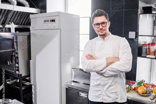 Portrait of confident young chef in eyeglasses standing in uniform with his arms crossed and looking at camera