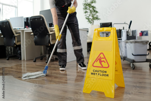 Yellow plastic signboard with caution warning about wet floor and female cleaner in black uniform working with mop in openspace office