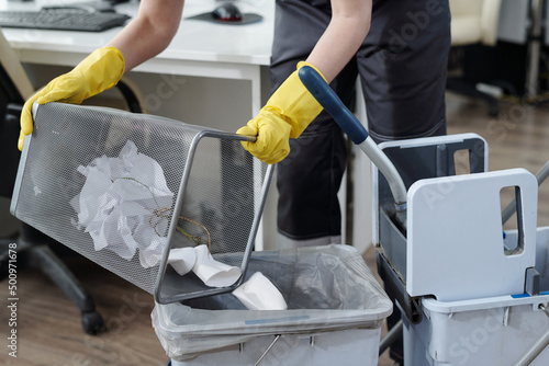 Gloved hands of female cleaner throwing trash from garbage bin into plastic bucket on janitor trolley while working in modern office photo