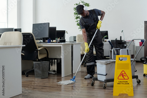 Contemporary young black man in workwear cleaning floor in openspace office in front of yellow plastic signboard with caution photo