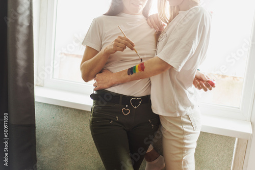 Lesbian couple hugs and painting together rainbow and holds brush. No face. Romantic embrace