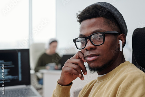 Face of young serious African American male programmer in eyeglasses, casualwear and earphones looking at camera by his workplace